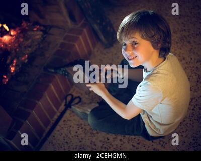 Fröhlicher junger Junge mit Balgen, der zu Hause am Kamin sitzt. Stockfoto