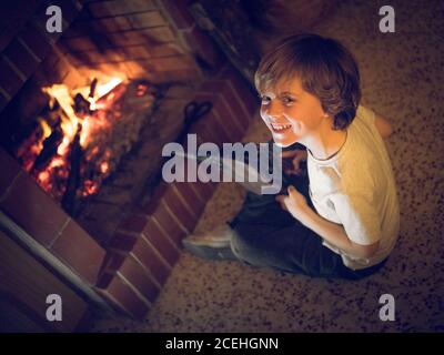 Fröhlicher junger Junge mit Balgen, der zu Hause am Kamin sitzt. Stockfoto