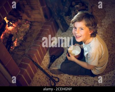 Fröhlicher junger Junge mit Balgen, der zu Hause am Kamin sitzt. Stockfoto