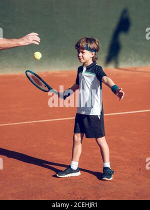 Crop Hand des alten Mannes in der Nähe des kleinen Kindes in Sportkleidung Mit Tennisschläger versucht, schwebenden Ball in der Luft zu treffen Auf dem Platz in sonnigen Tag Stockfoto