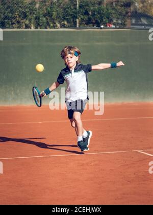 Kleines Kind in Sportbekleidung mit Tennisschläger versucht zu schlagen Schwebende Kugel in der Luft auf dem Platz an sonnigen Tag Stockfoto