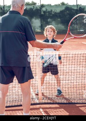 Zurück Ansicht Crop gealtertes Männchen und kleines Kind mit Tennis Schläger in der Nähe des Tennisnetzes auf dem Platz an sonnigen Tagen Stockfoto