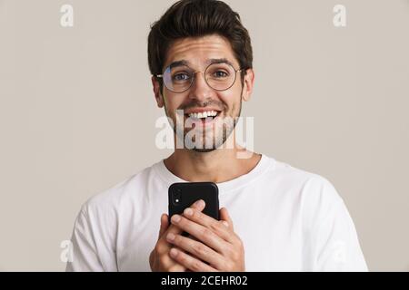 Bild von aufgeregt jungen Mann in Brillen halten Handy Isoliert durch beigefarbenen Hintergrund Stockfoto
