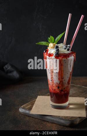 Köstlicher Beerenmilchshake garniert mit Schlagsahne, Heidelbeeren, tropfender Marmelade und Minze im Dunkeln. Stockfoto