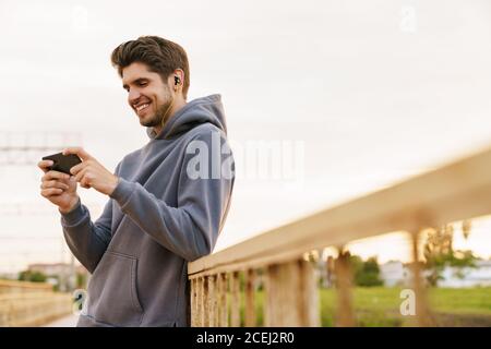 Bild von lächelndem Mann mit Kopfhörern, der Videospiel spielt Handy, während Sie sich im Freien auf dem Geländer lehnen Stockfoto