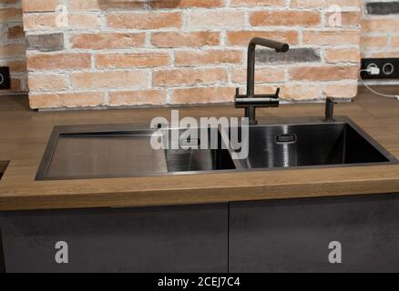 Moderne Küche mit schwarzem Waschbecken, Küchenarmatur für Wasser und Fronten vor dem Hintergrund eines Backsteins Wal. Braune und schwarze Holzarbeitsplatten. Luxus Stockfoto