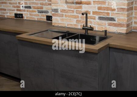 Moderne Küche mit schwarzem Waschbecken, Küchenarmatur für Wasser und Fronten vor dem Hintergrund eines Backsteins Wal. Braune und schwarze Holzarbeitsplatten. Luxus Stockfoto