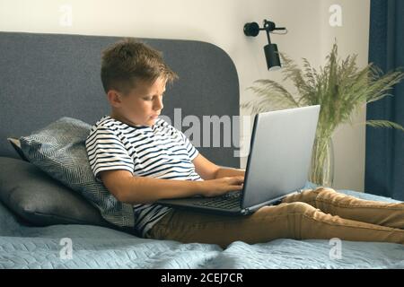 Junge mit seinem Laptop für Hausaufgaben, sitzt auf seinem Bett in modernen Innenraum. Online studieren während Quarantäne oder persönliche Bildungskonzept Stockfoto