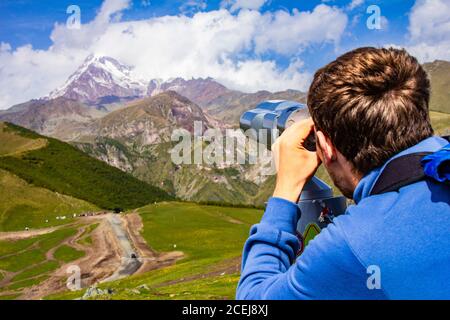 Mann, der im Binoskop auf die verschneiten Berggipfel gerichtet schaut Von Kazbeg aus der Sicht von Gergeti Trinity Church Tsminda Sameba Heilige Kirche in der Nähe Stockfoto