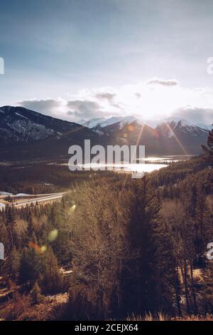 Helle Sonne scheint am Himmel mit wenigen Wolken über Herbst Kanadischer Wald im Hintergrund mit verschneiten Bergen und kleinem See Stockfoto