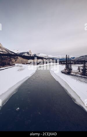 Fluss mit verschneiten Ufern, die durch Spalten fließen?in Winterwäldern mit Viele Tannen im Hintergrund mit Bergen und bewölktem Himmel Stockfoto