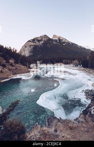 Fluss läuft in geschmolzenem Hochlandsee bei verschneiten Spalten mit Braune Felsen und einige Wintertannen an sonnigen Tagen Stockfoto