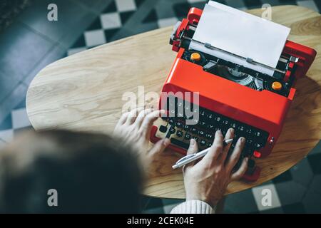 Crop Person halten Stift und Eingabe auf leuchtend rote Schreibmaschine An einem kleinen Tisch von oben gesehen Stockfoto