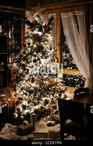 Wunderbarer Weihnachtsbaum mit verschiedenen Kugeln und helle Fee geschmückt Helle Girlande, die in der Nähe des Fensters in einem stilvollen Zimmer in der Nacht steht Stockfoto