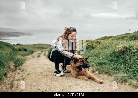 Ziemlich junge Frau lächeln und streicheln lustig Schäferhund während Verbringen Sie Zeit auf dem Land am bewölkten Tag Stockfoto
