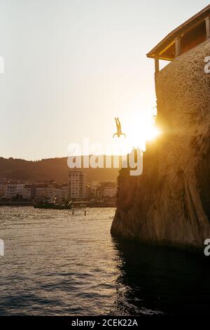 Silhouette des Mannes springt von der Klippe ins Meer auf dem Hintergrund Der Küstenstadt und Sonnenuntergang Sonne Stockfoto