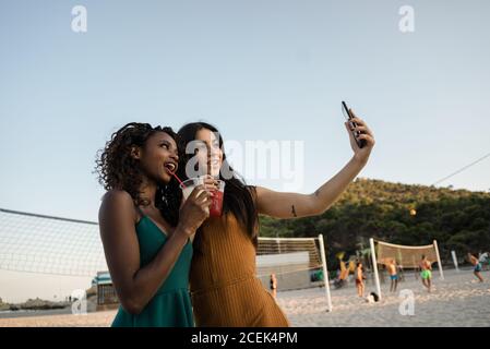 Stylische, vielfältige Frauen mit Getränken, die Selfie am Strand machen Stockfoto