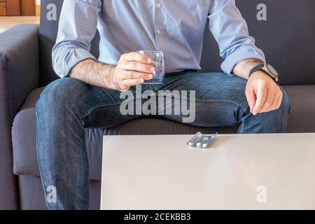 Der Mensch nimmt Pillen trinkt ein Glas Wasser und sitzt Auf dem Sofa in seinem Wohnzimmer Stockfoto