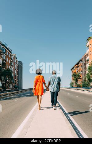Rückansicht des afroamerikanischen Paares in lässigen Outfits, die Hände halten Zusammen und zu Fuß entlang Bürgersteig zwischen zwei Straßen in der Stadt Stockfoto