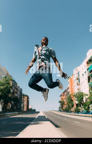 Hübscher afroamerikanischer Mann im lässigen Outfit, der rüber springt und schreit Straße in der schönen Stadt an sonnigen Tag Stockfoto