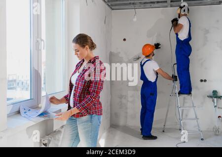 Zwei Arbeiter in Overalls und schalldichte Kopfhörer bohren die Wand. Der Kunde prüft die Aufteilung der Wohnung Stockfoto