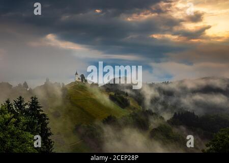 Jamnik, Slowenien - magischer nebliger Sommeraufgang an der Kirche Jamnik St.Primoz. Der Nebel geht sanft an der kleinen Kapelle mit buntem Himmel und den Julischen Alpen vorbei Stockfoto