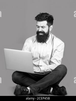 Lächelnd schöner bärtiger Mann Arbeiter Laptop. Glücklicher junger Mann, der auf dem Boden mit und mit Laptop-Computer auf rotem Hintergrund sitzt. Junger Geschäftsmann Stockfoto