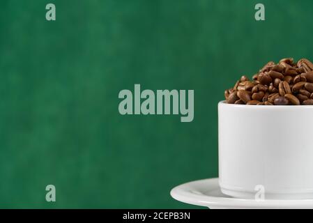 Internationaler Tag des Kaffees Konzept. Nahaufnahme weiße Kaffeetasse voller Kaffeebohnen auf dunkelgrünem Hintergrund. Stockfoto