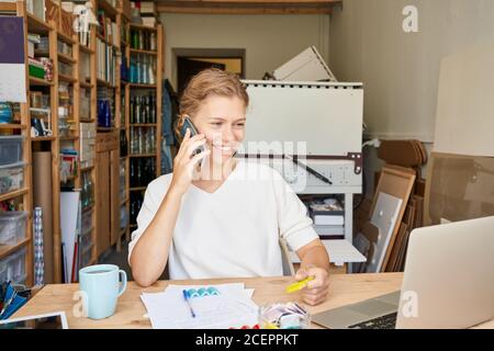 Junge Unternehmerin Designerin im Gespräch auf Handy und Blick auf Laptop und lächelnd, glückliche Dame in sonnigen Tag in der Werkstatt. Kleine Unternehmen für Frauen Stockfoto