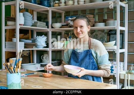 Qualifizierte junge Frau in Schürze am Tisch stehend und Zeichnung auf Keramik-Schüssel in Töpferei. Erwachsene Kunsthandwerkerin mit Blick auf die Kamera und lächelnd Stockfoto