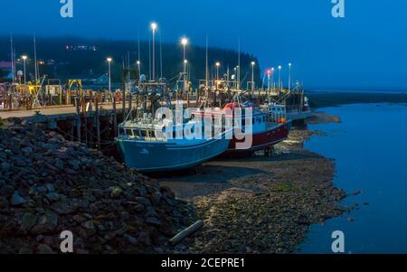 Einbruch der Dunkelheit über dem nebligen Hafen. Fischerboote vertäuten am Dock und ruhten bei Ebbe auf dem Meeresboden. Bay of Fundy, Alma Harbor, New Brunswick, Kanada. Stockfoto