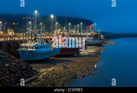 Einbruch der Dunkelheit über dem nebligen Hafen. Fischerboote vertäuten am Dock und ruhten bei Ebbe auf dem Meeresboden. Bay of Fundy, Alma Harbor, New Brunswick, Kanada. Stockfoto