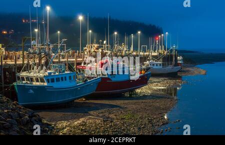 Einbruch der Dunkelheit über dem nebligen Hafen. Fischerboote vertäuten am Dock und ruhten bei Ebbe auf dem Meeresboden. Bay of Fundy, Alma Harbor, New Brunswick, Kanada. Stockfoto