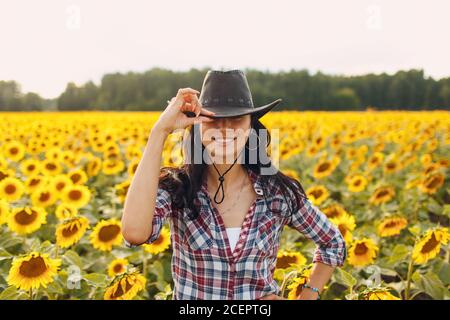 Junge Frau Agronomin trägt Cowboyhut, kariertes Hemd und Jeans auf Sonnenblumenfeld. Umfassendes Erntekonzept. Stockfoto