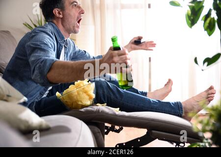 Wütender Mann, der Sport im Fernsehen sieht, sitzt bequem auf einem Sofa und hat einen Snack mit Bier und einer Fernbedienung in den Händen. Stockfoto