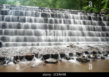 Vollformat-Langzeitbelichtung eines mehrstufigen Wasserfalls in einem Waldwasserstraßkanal mit grünem Laub hinten und glattem Wasser im Vordergrund. Stockfoto