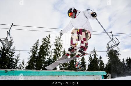 Männlicher Skifahrer in Skijacke und Helm macht Sprung am Wintertag. Mann Freerider springen in der Luft im Skigebiet mit Pinien und Skilifte im Hintergrund. Konzept von Wintersport und aktiver Freizeit. Stockfoto