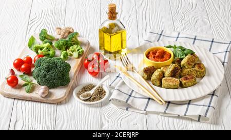 Gebackene Brokkoli auf einem weißen Teller mit Marinara-Sauce auf einem Holztisch mit Zutaten auf einem Schneidebrett und goldenem Besteck, horizontale Ansicht von Stockfoto