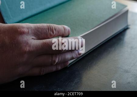 Eine reife männliche Hand öffnet ein Buch mit einer grünen ersten Seite. Dickes Hardcover-Buch. Männliche Hand mit einem Buch auf einem schwarzen Tisch. Selektiver Fokus. Stockfoto