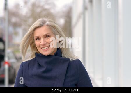 Selbstbewusst stilvolle freundliche Frau mittleren Alters zu Fuß durch eine Stadt Straße mit der Brise weht ihr langes Haar, wie sie Lächelt die Kamera an Stockfoto