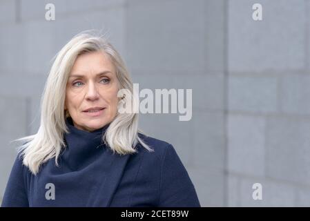 Nachdenklich attraktive blonde Frau beiseite, als sie draußen in einem warmen blauen Wintermantel gegen eine graue Wand Hintergrund steht Stockfoto