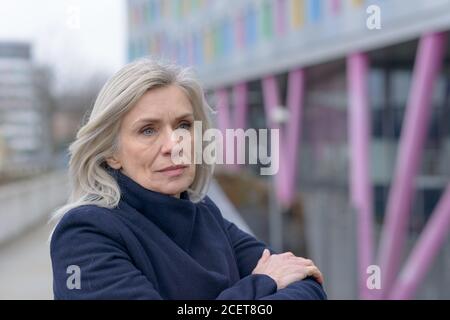 Nachdenklich reife attraktive blonde Frau Blick zurück über die Schulter zu, wie sie sich in Anlehnung an den Rand Geländer einer städtischen Außen walkwa Stockfoto