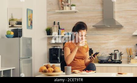 Frau stöbert beim Frühstück am Telefon und trinkt frischen Orangensaft mit einer gesunden Bio-Mahlzeit und einer Tasse Kaffee, mit moderner Technologie allein in der Küche, bevor sie zur Arbeit geht. Stockfoto