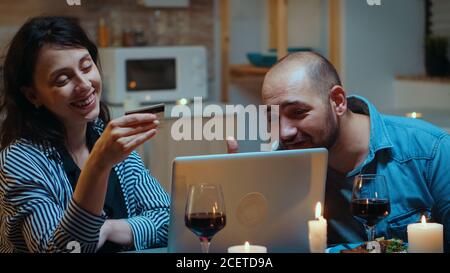 Ehemann und Ehefrau kaufen online ein und zahlen während des Abendessens mit Karte. Pärchen sitzen am Tisch, suchen, surfen, surfen, Technologie nutzen, festliches Essen im Speisesaal genießen Stockfoto