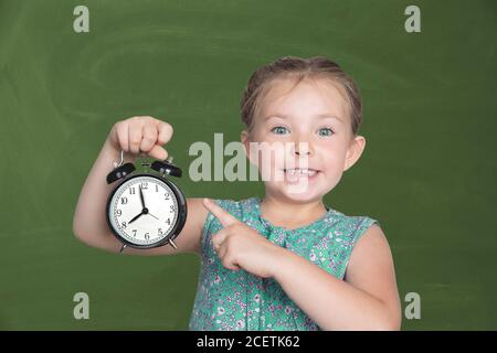 Ein kleines Mädchen hält einen Wecker gegen die grüne Schulbank. 8 Uhr Es ist Zeit für die Schule. Hochwertige Fotos Stockfoto