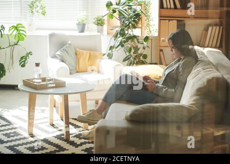 Horizontal durch Glas Schuss von schönen jungen Frau entspannend auf Sofa Surfen Internet auf digitalem Tablet Stockfoto
