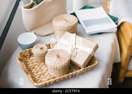 Ein Geschenkkarton mit beigefarbenem Satinband mit Schleife, Strohkörben mit Deckel auf einem Korbtablett und einem Becher. Komfort zu Hause Stockfoto