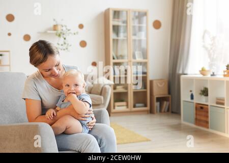 Horizontales Porträt einer glücklichen jungen Frau auf dem Sessel sitzend mit ihrem kleinen Kind auf dem Schoß lächelnd, Kopierraum Stockfoto