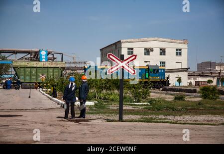 Taraz/Kasachstan - April 25 2012: Phosphordüngeanlage. Zwei Arbeiter warten auf den Zug. Blaue Lokomotive und grüner Eisenbahnwagen auf Industrie b Stockfoto