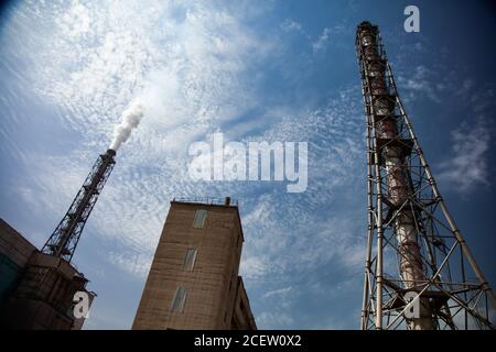 Fabrikschornsteine mit Trägern und Industriegebäude am blauen Himmel mit Rauch und hellweißen Wolken. Panoramaansicht. Stockfoto
